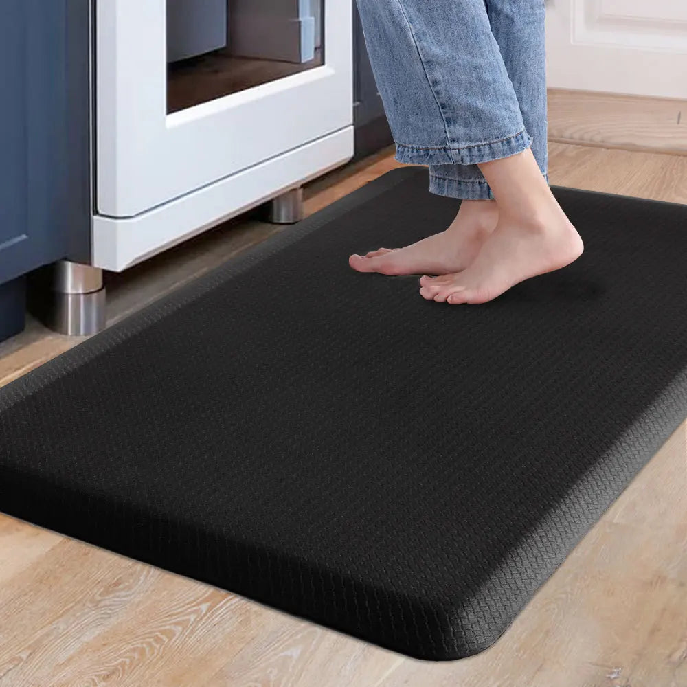 A person's feet are standing on a black anti-fatigue PVC kitchen mat placed on a light wooden floor near a white appliance.