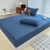 A fitted bed sheet cover in a solid blue color is placed on a bed, with a teddy bear and some books nearby.