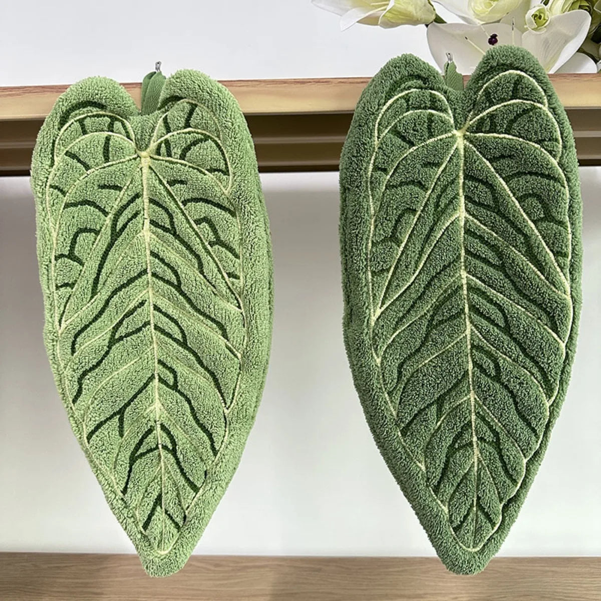 Two green plant design hand towels with textured leaves displayed on a wooden shelf.