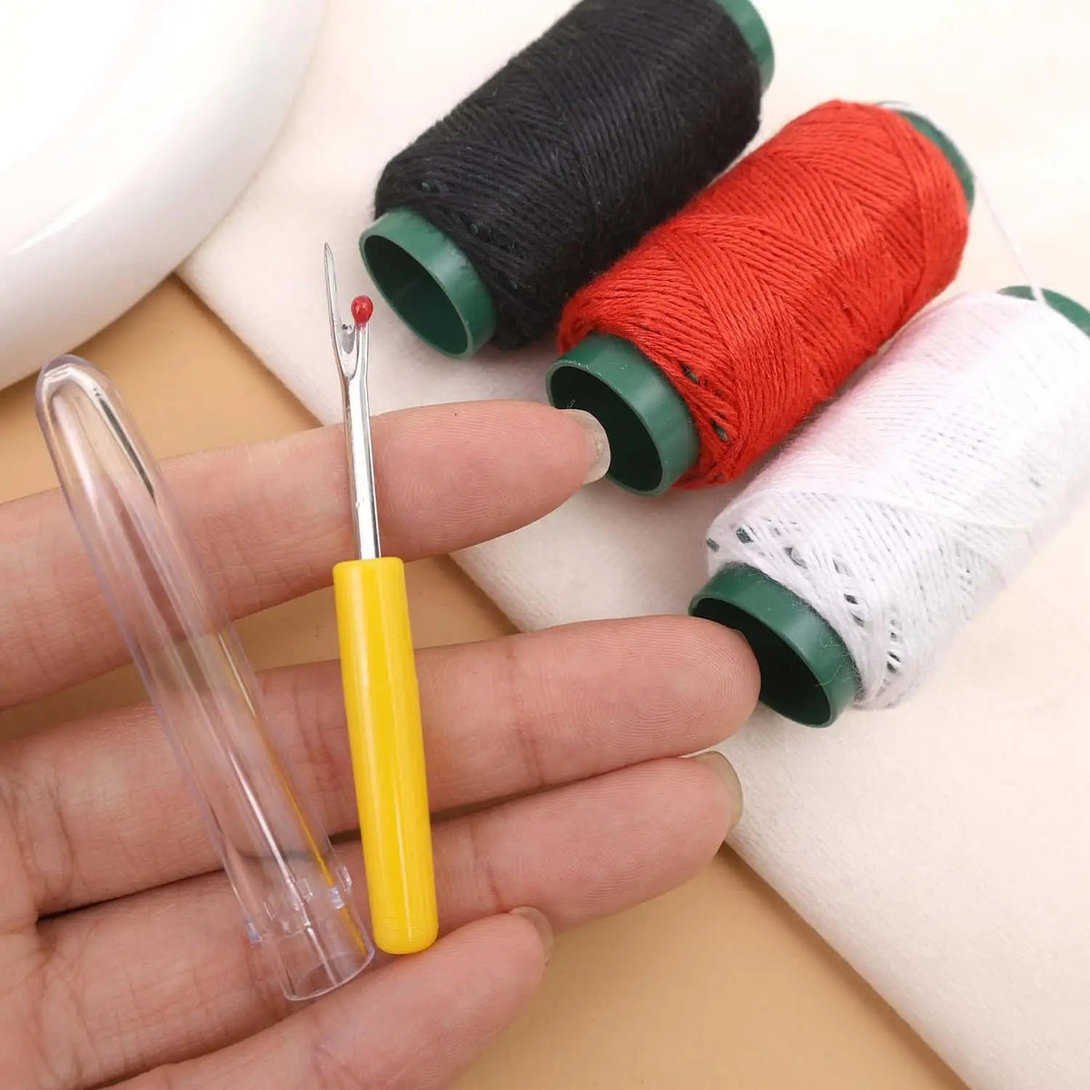 A hand holds a pair of sewing needles and several spools of colored embroidery thread for a heavy duty leather kit.