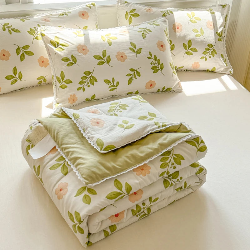 A stack of white quilted blankets with green and pink floral patterns and matching pillows on a light surface.
