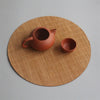 A terracotta teapot and bowl resting on a round, woven bamboo tea mat.