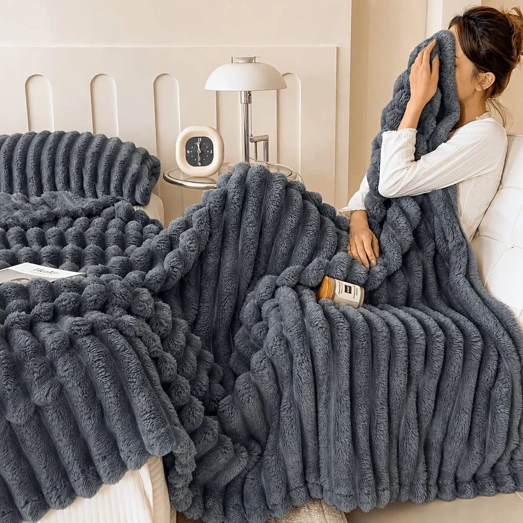 A dark gray, chunky fleece throw blanket is draped over a white sofa next to a lamp.