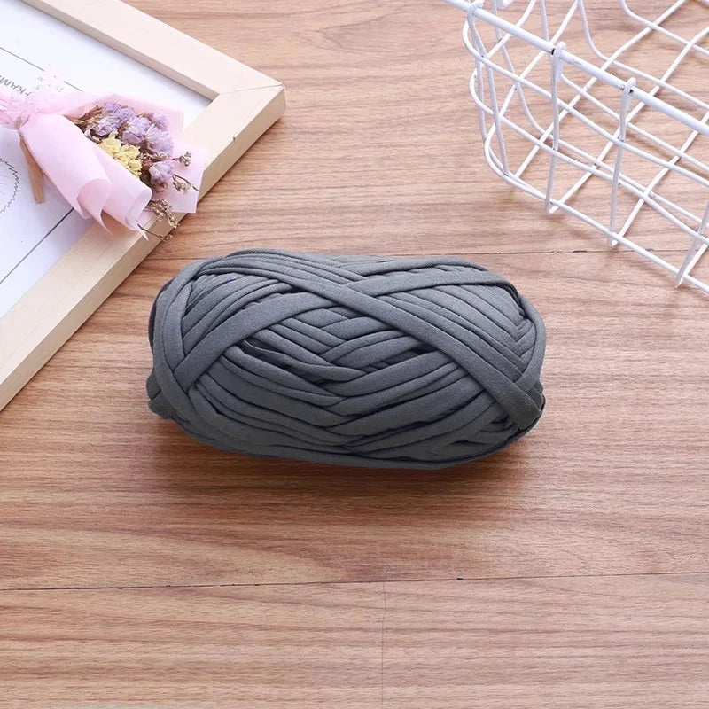 A ball of gray flat thread yarn lying on a wooden surface next to a white wire basket and some pink flowers.