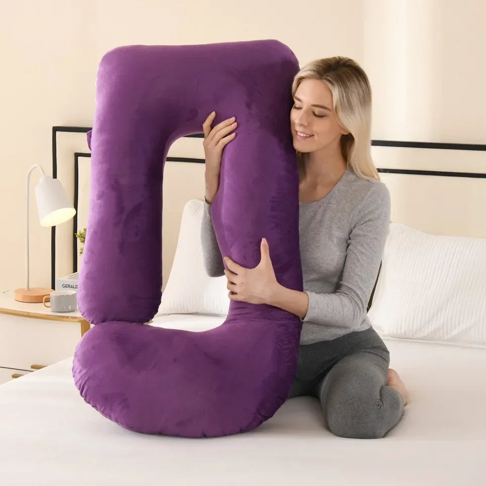 A woman is holding a purple J-shaped ergonomic maternity pillow on her bed.