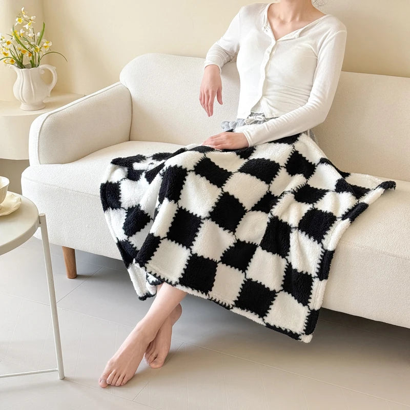 A woman wearing a white top and skirt sits on a sofa with a black and white checkered fleece blanket draped over her lap.