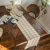 A rustic wooden table is set with a beige and white patterned linen table runner, books, and greenery near a window.