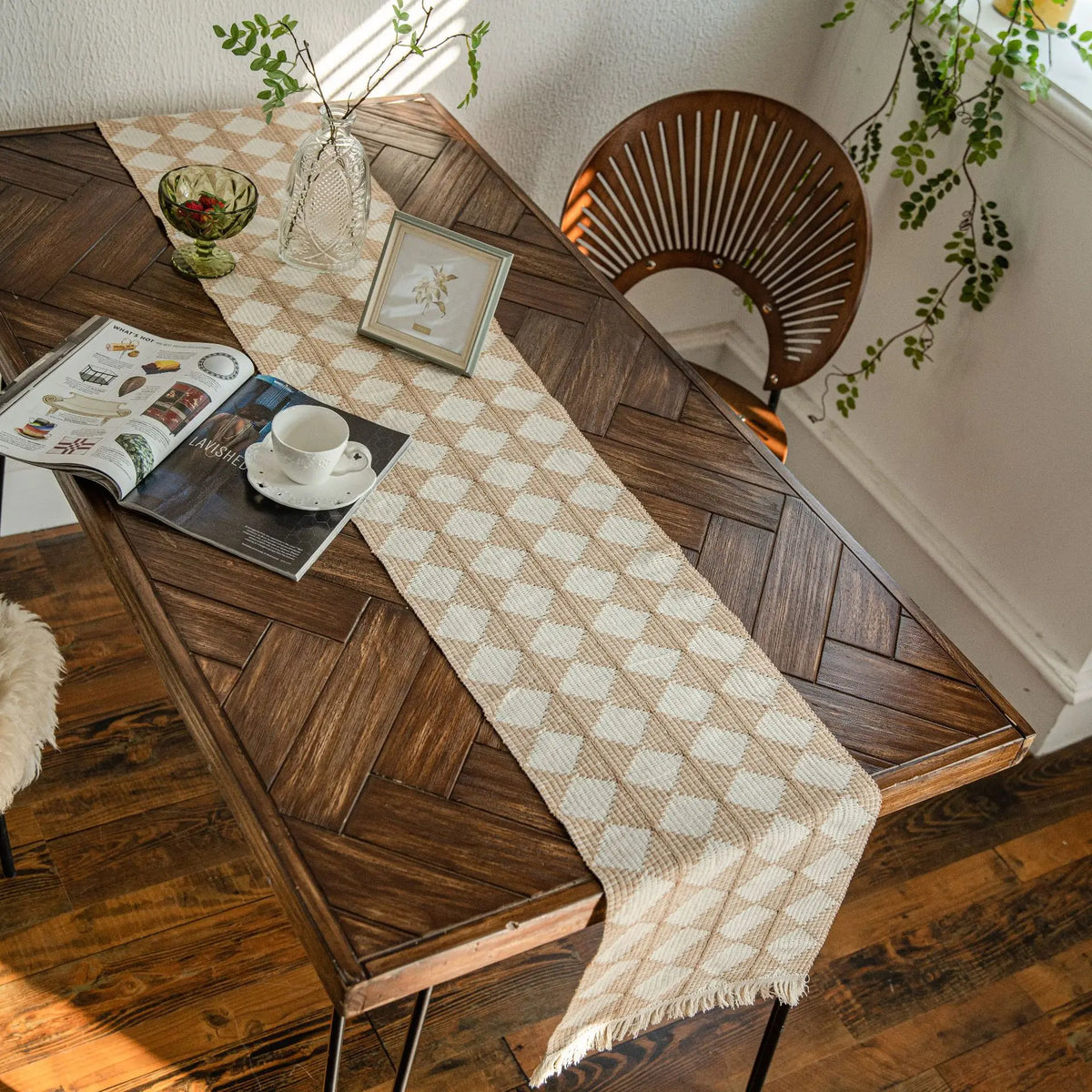 A rustic wooden table is set with a beige and white patterned linen table runner, books, and greenery near a window.