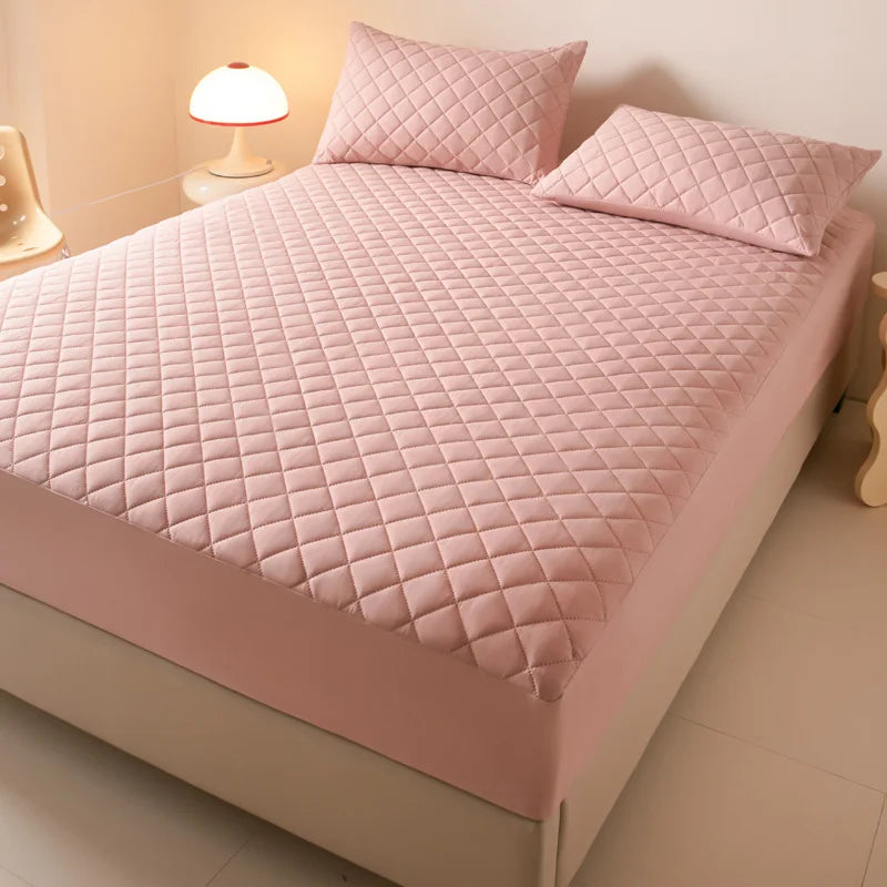 A pink quilted mattress protector is displayed on a bed with matching pillows and a bedside lamp.