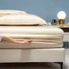 A hand adjusts the smooth, beige luxury bamboo bedding on a bed against a light blue wall.