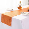A golden sequin table runner laid on a white tablecloth with silverware and a bottle of liquor.