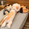 A large, light-colored dog plush pillow doll is lying down on a table covered with a gray cloth.
