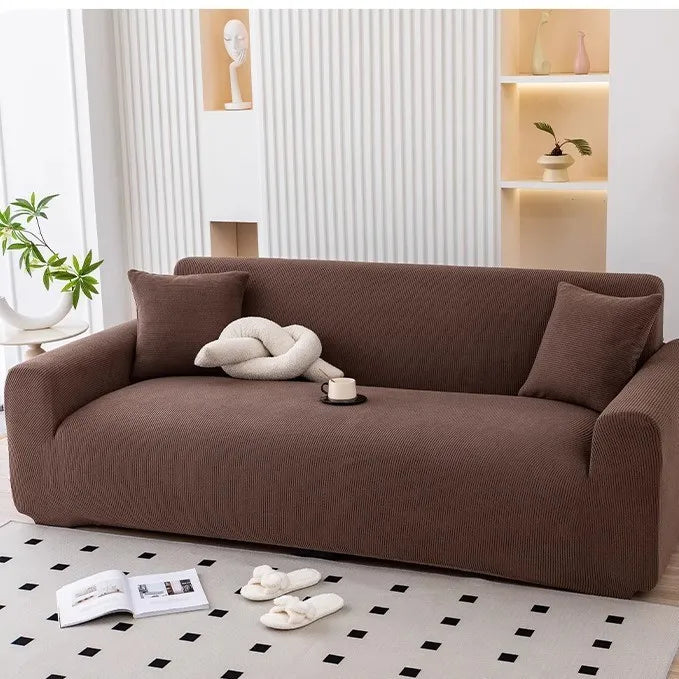 A brown stretchable full-wrap sofa slipcover is shown on a light rug with white slippers nearby.