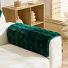 A white sofa cushion is draped with a deep emerald green faux fur throw on a light wood shelf.