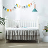 A white crib with a ruffled gray bed skirt and a teal pendant light hanging above it in a bright nursery room.