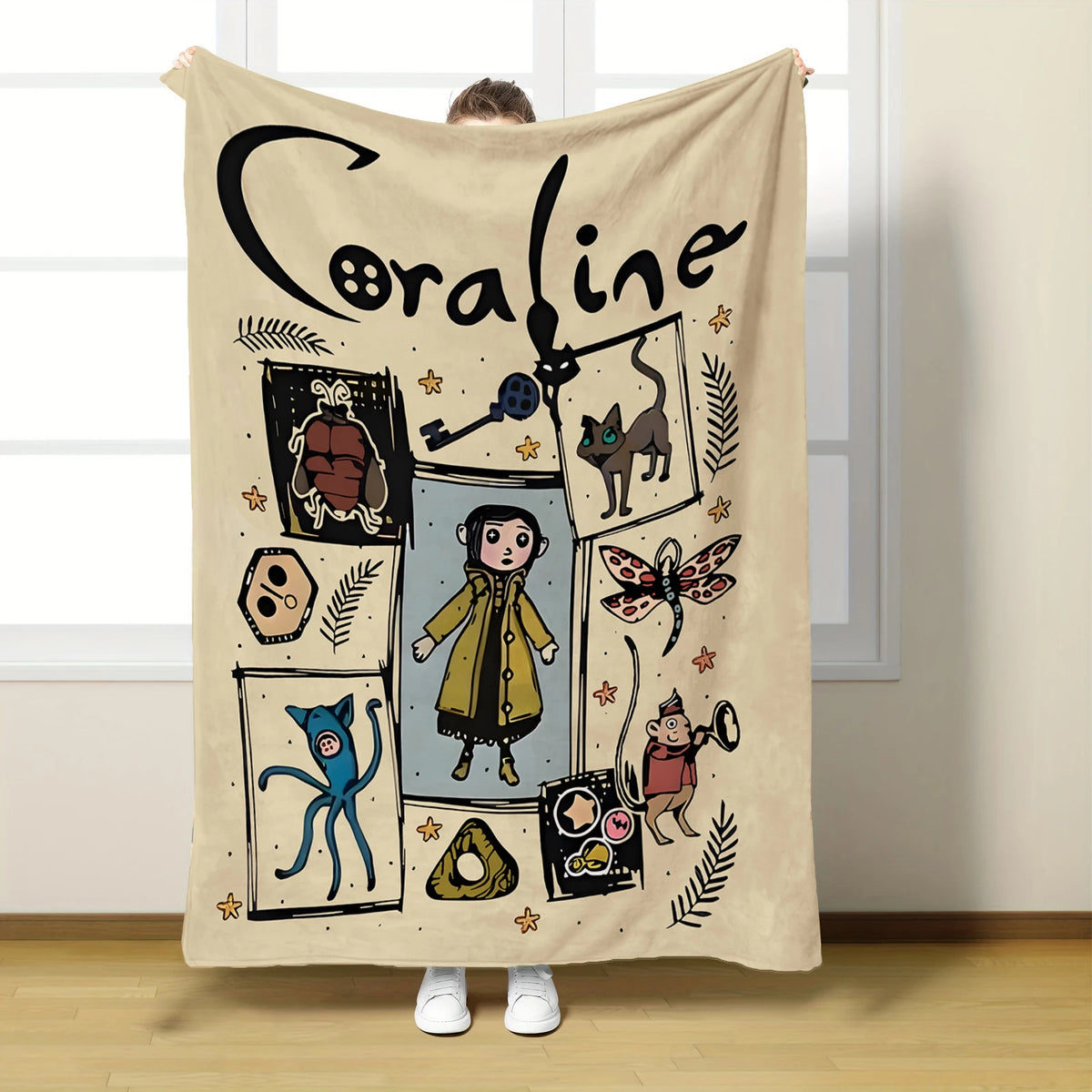 A beige flannel throw blanket featuring a black line drawing of a girl named Coraline surrounded by various whimsical illustr