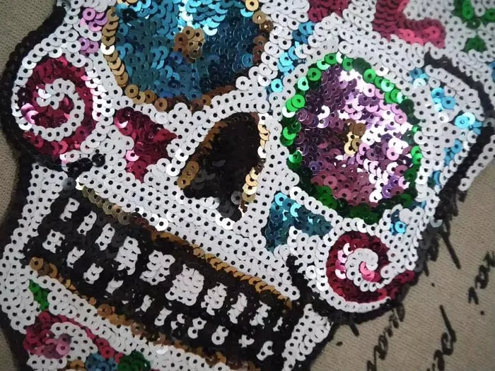 A close-up of a sequin skull embroidered clothing patch featuring purple, teal, and pink sequins.