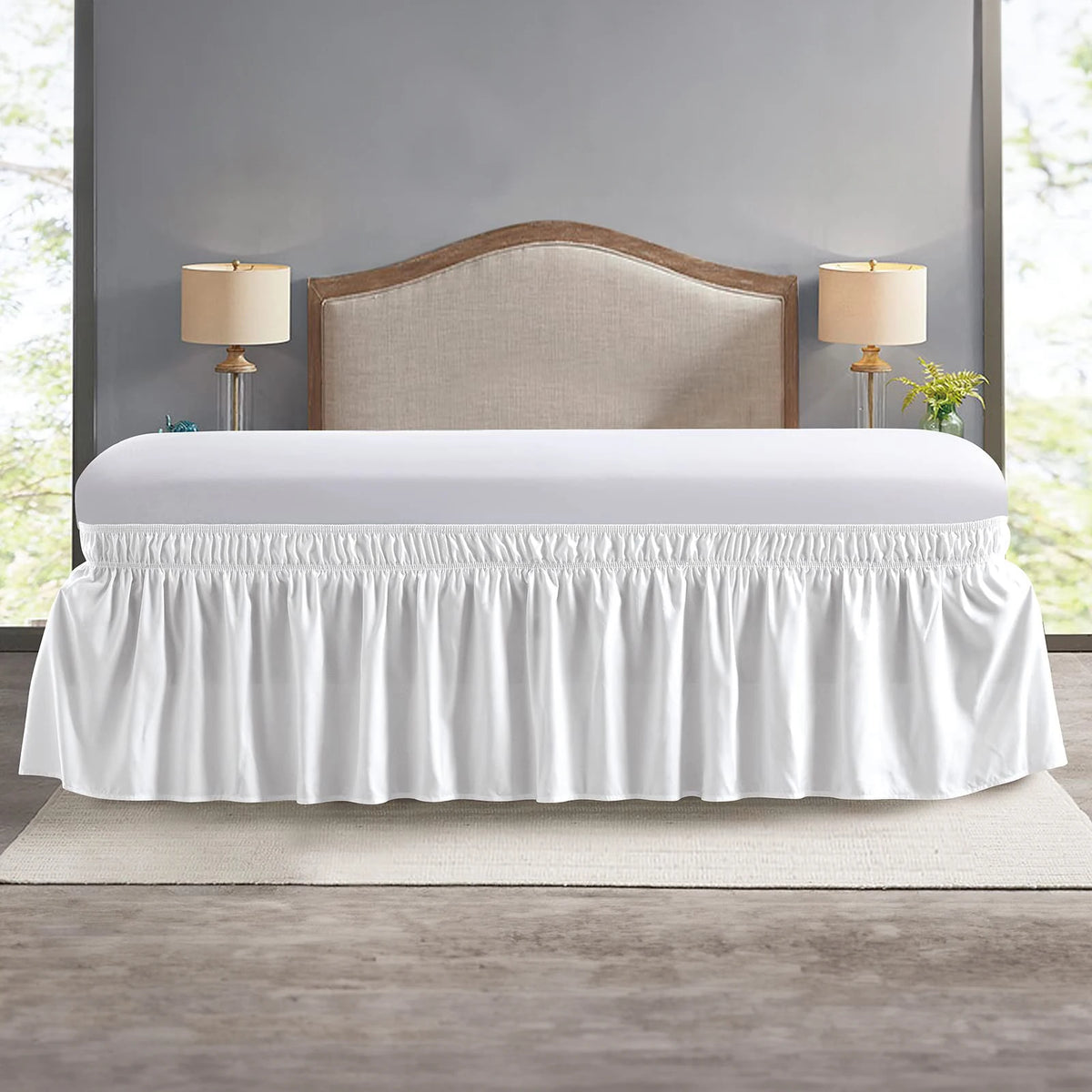 Elegant white elastic bed skirt with ruffles displayed on a bed against a gray wall.