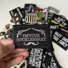 A hand holds a black iron-on slogan patch featuring the text I'm Your Huckleberry among other similar patches.