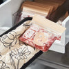 A clear plastic bag of pinkish-red floral heat-resistant oven mitts is placed on a quilted black holder.