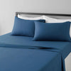 A set of four blue microfiber flat fitted sheets on a bed with white pillows and a dark headboard.