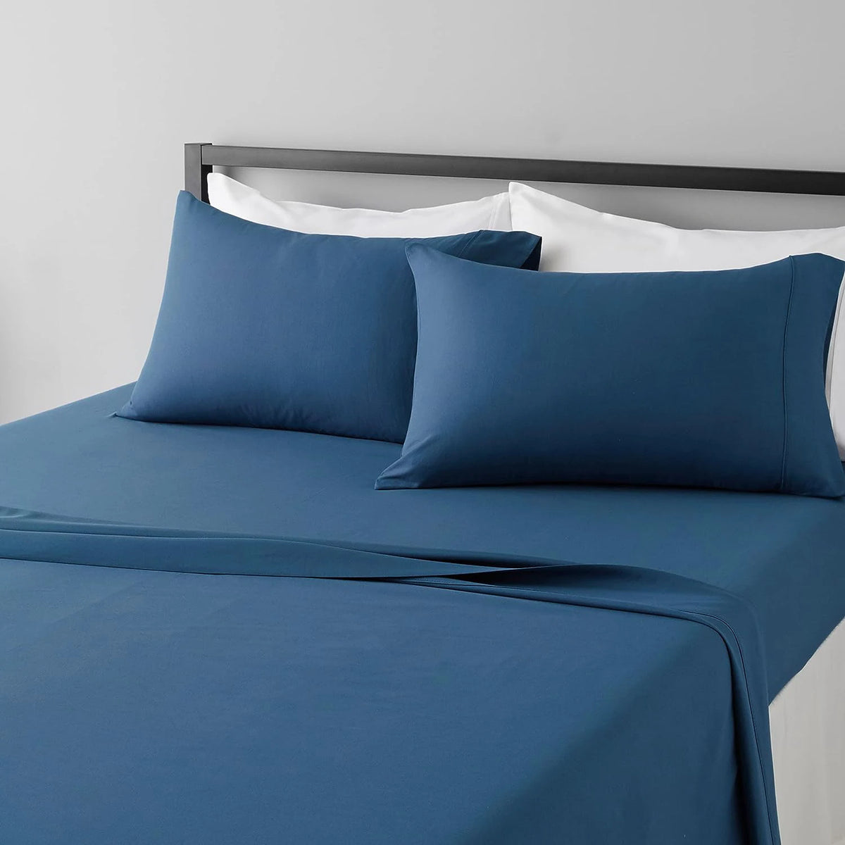 A set of four blue microfiber flat fitted sheets on a bed with white pillows and a dark headboard.