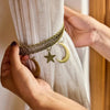 A hand is holding a gold-toned braided clip with a star and a crescent moon charm against a light fabric curtain.
