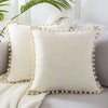 A white velvet decorative cushion cover with a ruffled trim rests on a gray sofa next to a knitted throw.