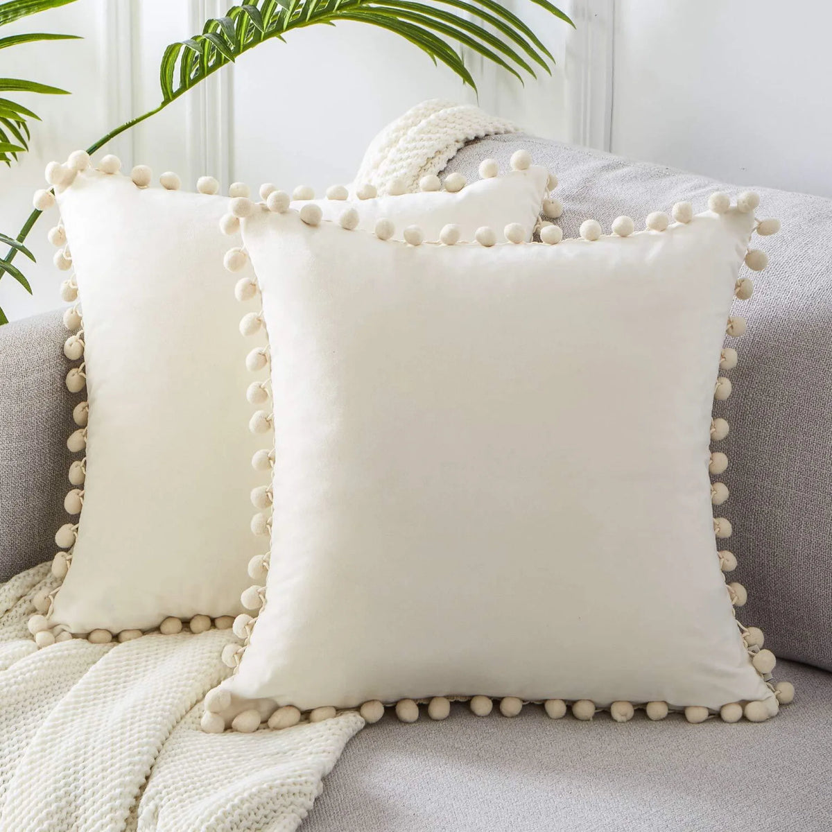 A white velvet decorative cushion cover with a ruffled trim rests on a gray sofa next to a knitted throw.