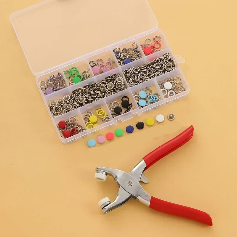 A clear plastic box filled with various colorful metal sewing buttons and a pair of silver-handled pliers.