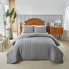 A light gray quilted bedspread set is displayed on a bed with a wooden headboard and white paneling.