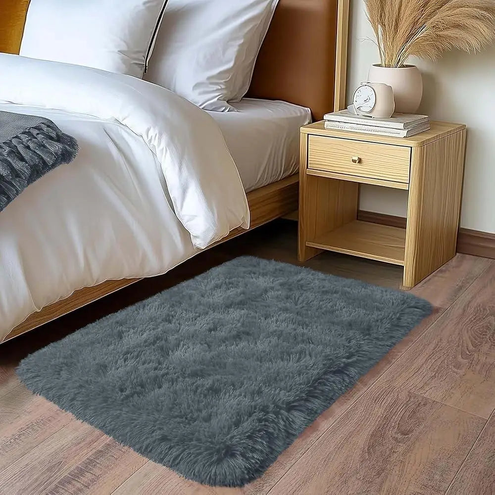 A plush gray area rug is placed on a wooden floor next to a bed and a bedside table.