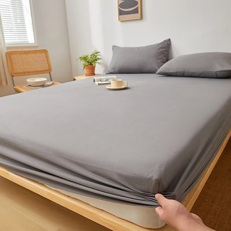 A gray fitted sheet is being folded on a wooden bed frame, showing its deep pockets and elastic edges.