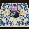 A mat featuring a central iridescent sphere surrounded by blue flowers and celestial elements for tarot readings.