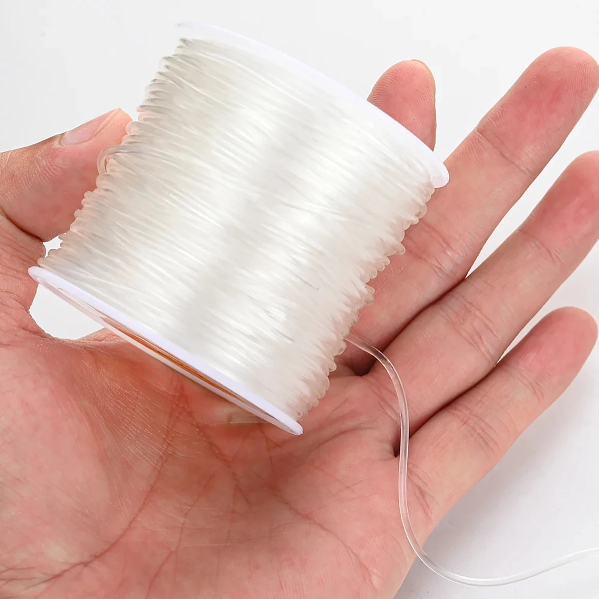 A hand holds a spool of transparent elastic cord used for jewelry making.