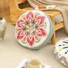 A round cushion with a colorful, mandala-like floral pattern is placed on a wooden floor frame.