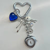 Silver keychain featuring a heart charm, a blue gemstone, and a pocket watch.