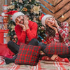 A smiling woman in a red sweater and plaid pants is relaxing on a rug next to a decorated Christmas tree.