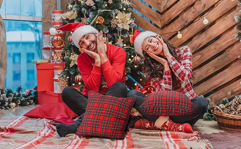 A smiling woman in a red sweater and plaid pants is relaxing on a rug next to a decorated Christmas tree.