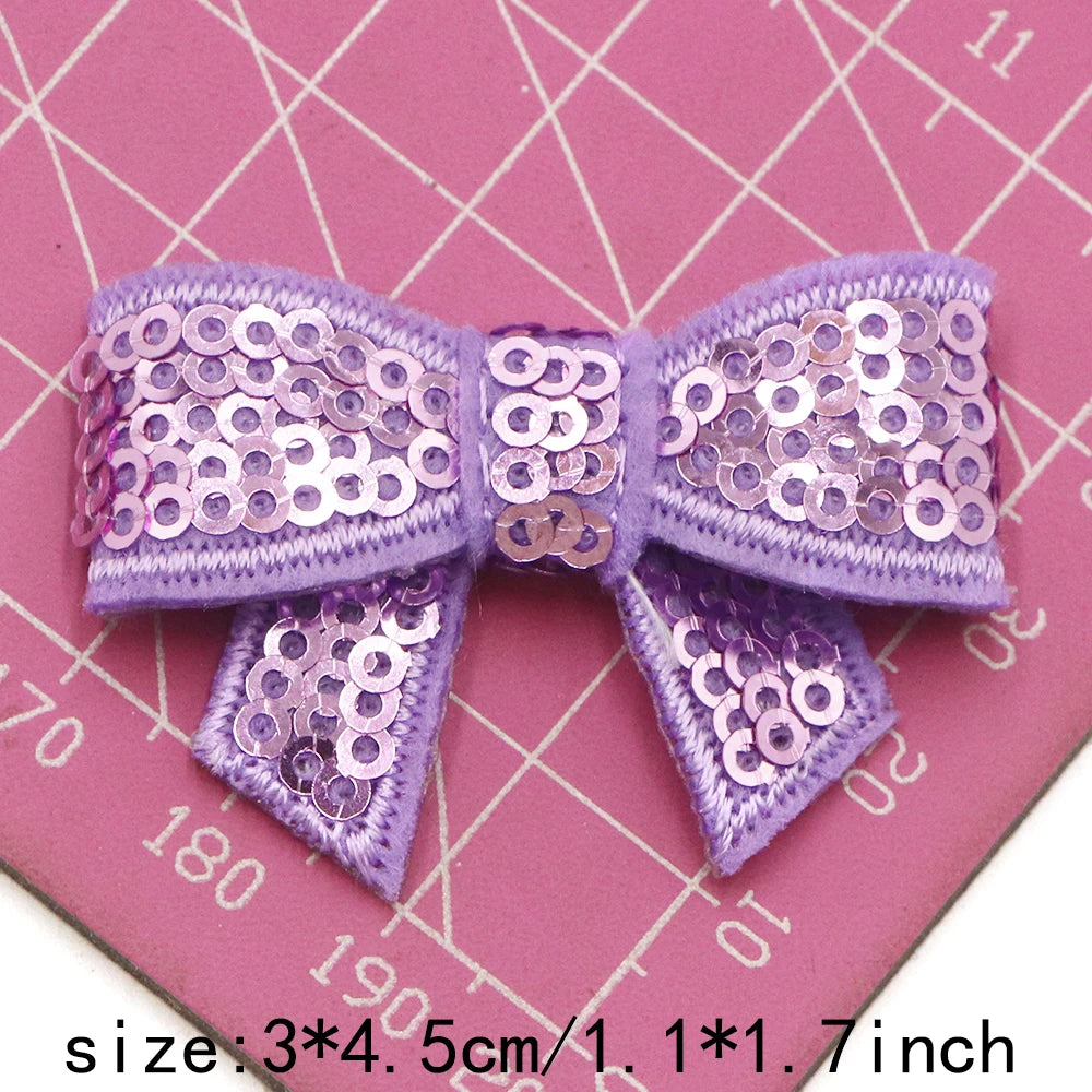 Purple sequin bow patches for DIY decoration shown on a pink grid background