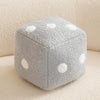A gray plush dice cube cushion with white polka dots resting on a light beige surface.