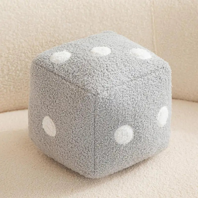 A gray plush dice cube cushion with white polka dots resting on a light beige surface.