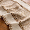 A beige linen table runner with macrame tassels and fringed edges laid on a wooden surface.