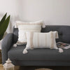 A white knitted throw pillow cover with beige tassels resting on a gray sofa.