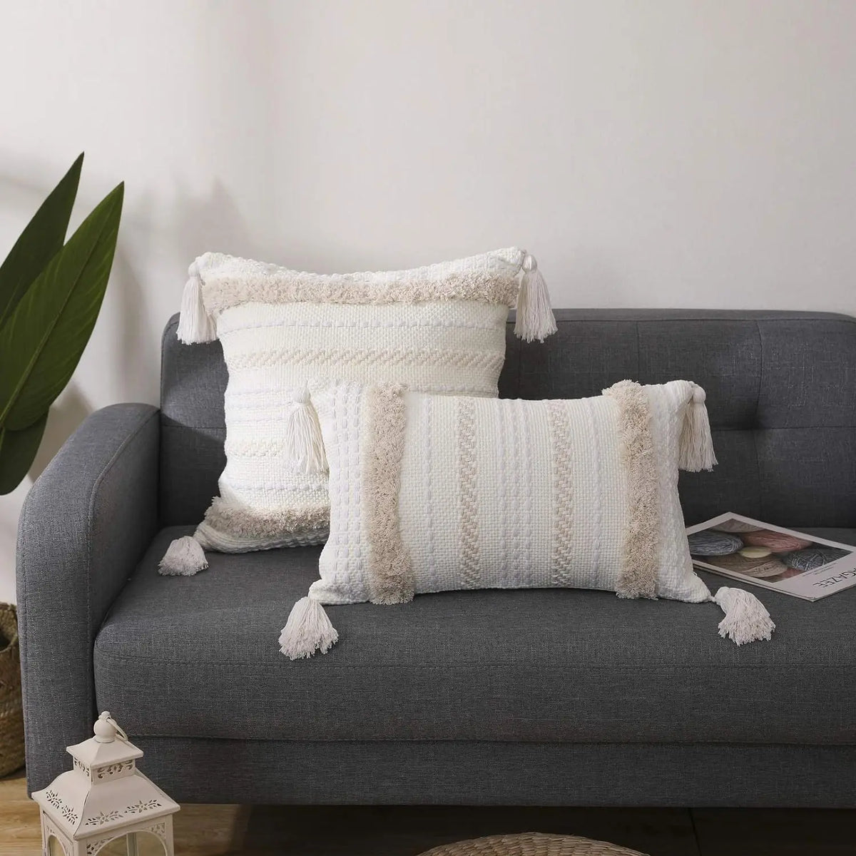 A white knitted throw pillow cover with beige tassels resting on a gray sofa.