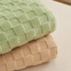 A stack of cozy checkered fleece throw blankets in sage green and beige tones.