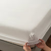 A hand is adjusting the elastic edge of a white soft touch microfiber fitted sheet on a bed.
