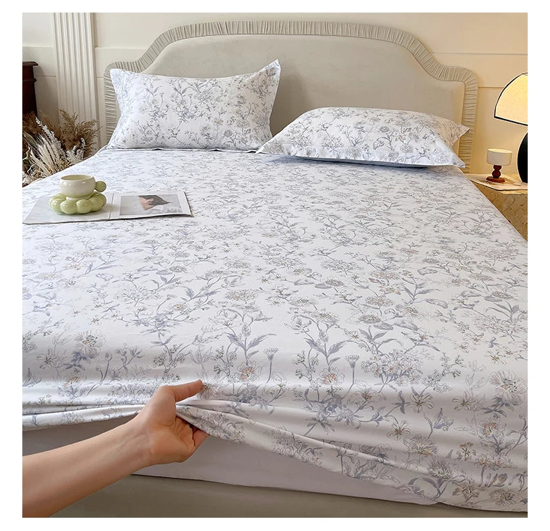 A white floral printed cotton fitted sheet is displayed on a bed with a upholstered headboard.