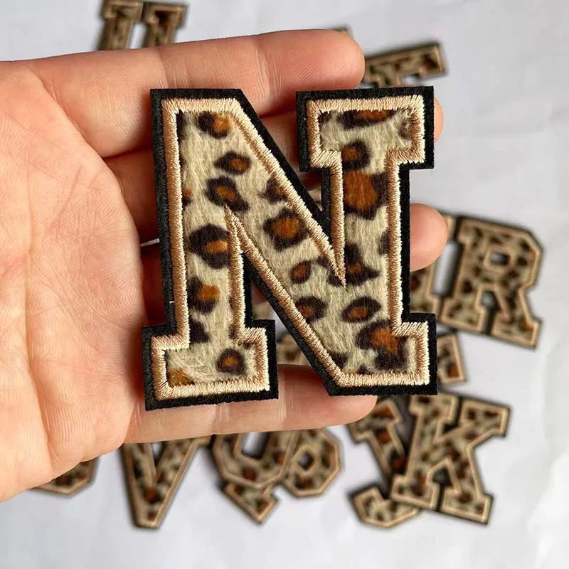 A hand holds a leopard print alphabet iron-on patch letter N and several other letters.