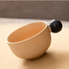 A small, beige ceramic cup with a black handle resting on a light yellow textured surface.